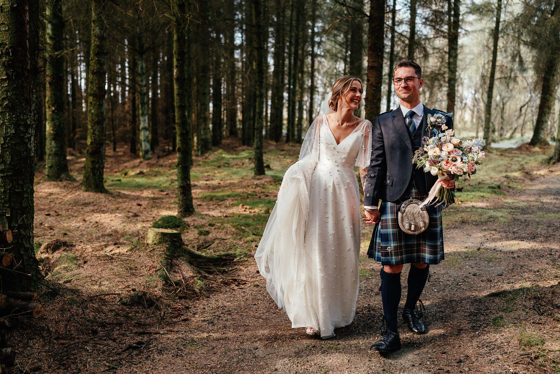 Bride and groom walking through the woods smiling with flowers